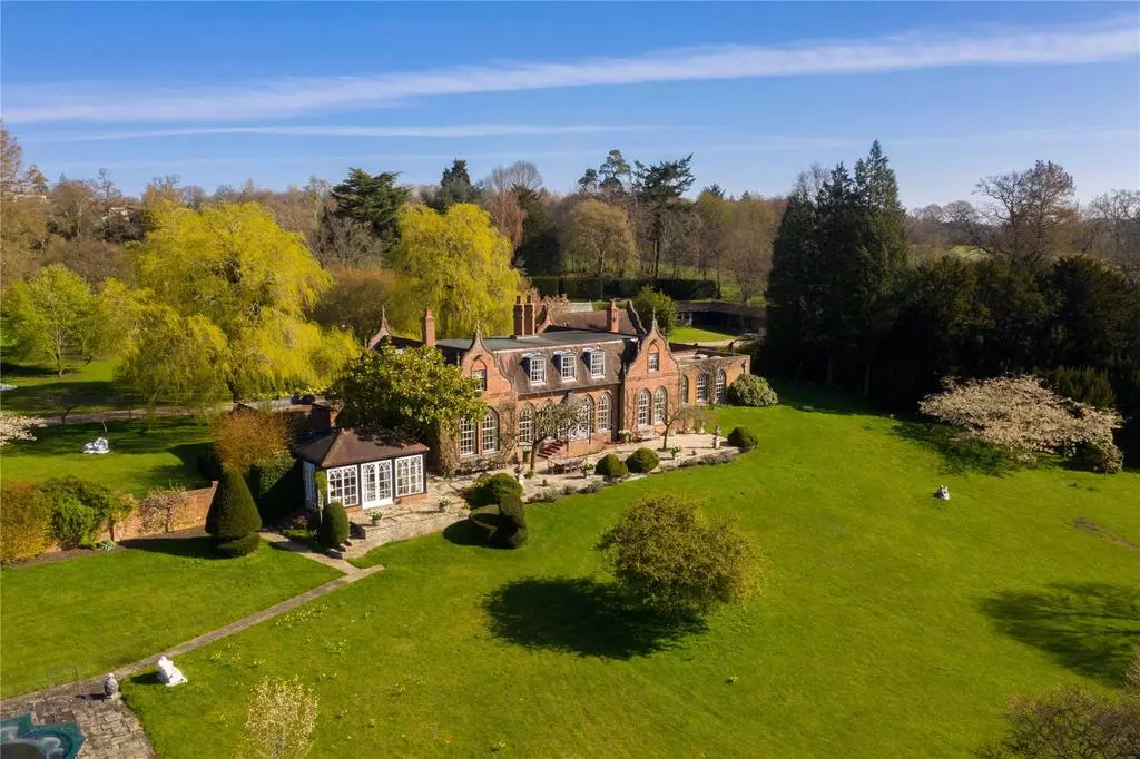 Large red-brick country house with arched windows and chimneys set in spacious green gardens with trees under a blue sky.