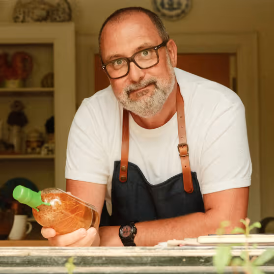 Bearded man wearing glasses and a dark apron holds a bottle with a green cap, leaning forward across a counter in a cozy kitchen.