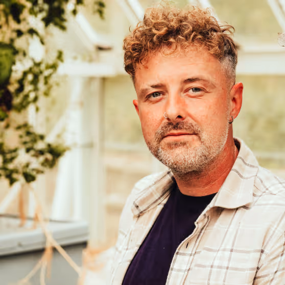 Man with curly light brown hair, nose ring, and beard wearing a checkered shirt over a dark t-shirt indoors near plants.