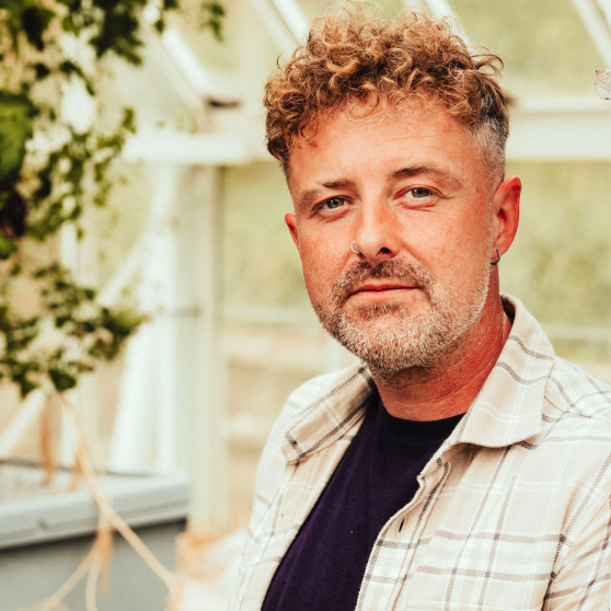Man with curly light brown hair, nose ring, and beard wearing a checkered shirt over a dark t-shirt indoors near plants.