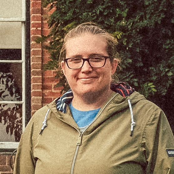 Person wearing glasses and a green jacket smiling outdoors with brick wall and greenery in background.