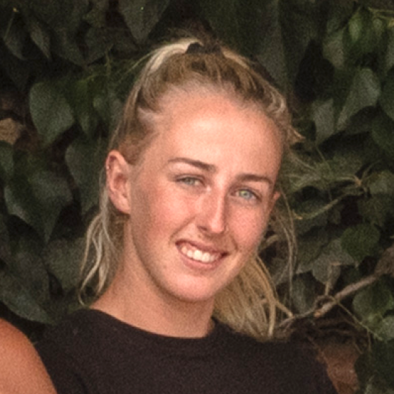 Young woman with blonde hair tied back smiling against a leafy green background.