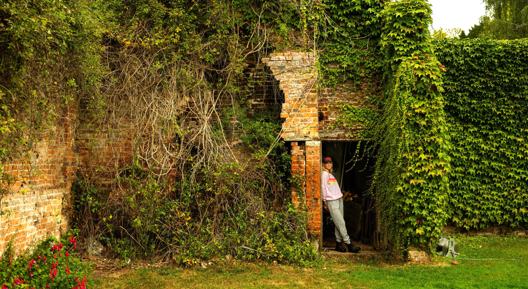 Person in casual clothes leaning against the doorway of an old brick building overgrown with ivy and vegetation.