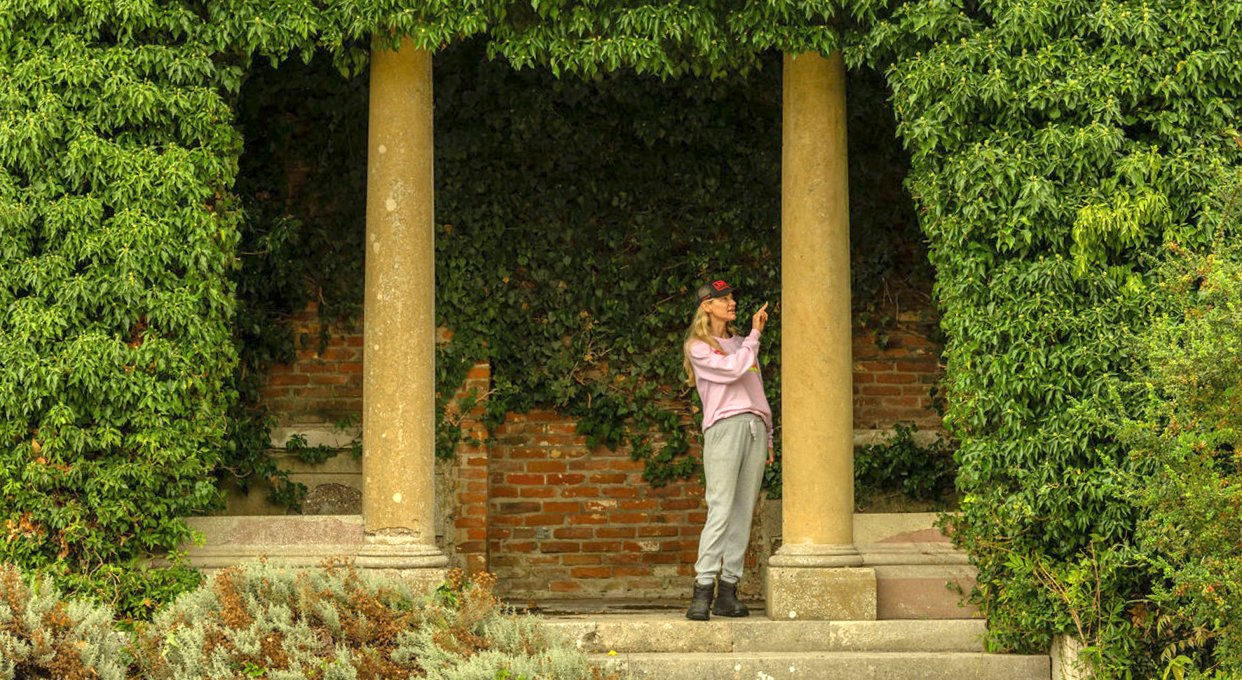 Woman in casual clothes pointing upwards while standing between two stone columns covered with green ivy.