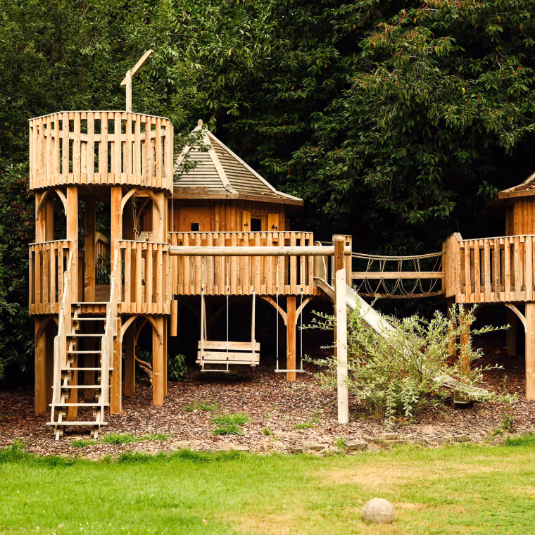Wooden playground structure with towers, bridge, slide, and a swing, set in a garden with green grass and dense trees in the background.