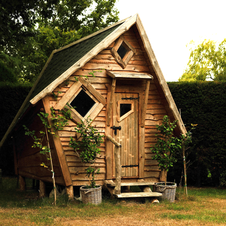 Small wooden garden hut with a triangular roof, diamond-shaped windows, and two potted plants beside the entrance.