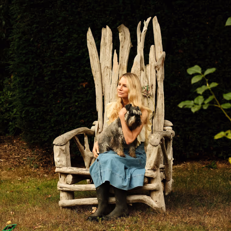 Blonde woman in a denim skirt and black boots sitting on a rustic wooden chair outdoors holding a small grey dog.