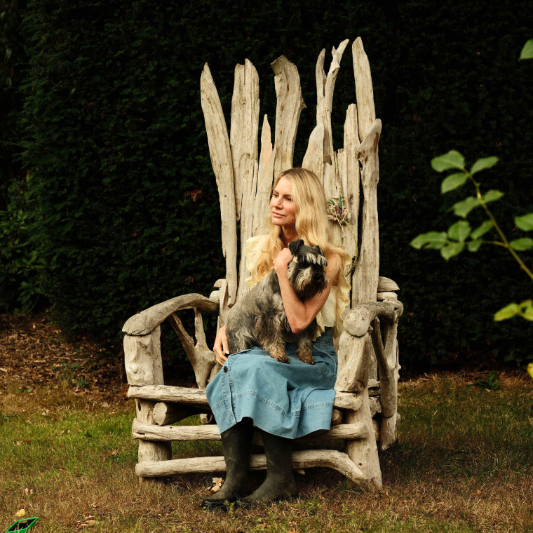 Blonde woman in a denim skirt and black boots sitting on a rustic wooden chair outdoors holding a small grey dog.