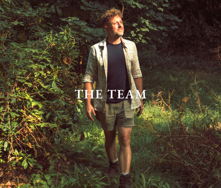Man with curly hair walking on a forest path surrounded by green foliage, lit by warm sunlight.