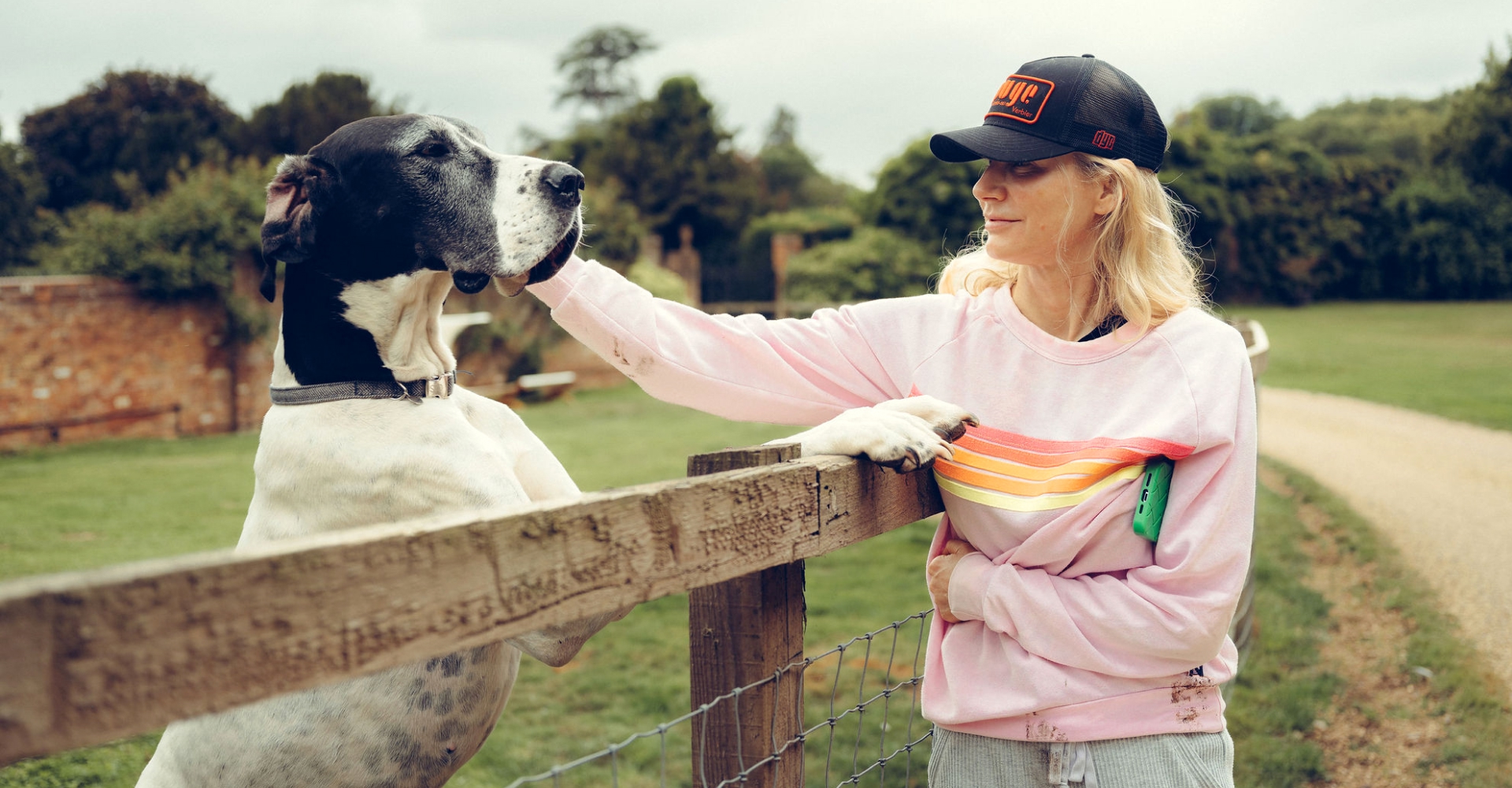 Woman in a pink sweatshirt and cap petting a large black and white dog over a wooden fence in a green park.
