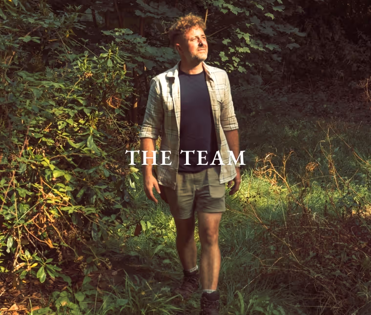 Man with curly hair wearing a checked shirt and shorts walking through a sunlit forest path.