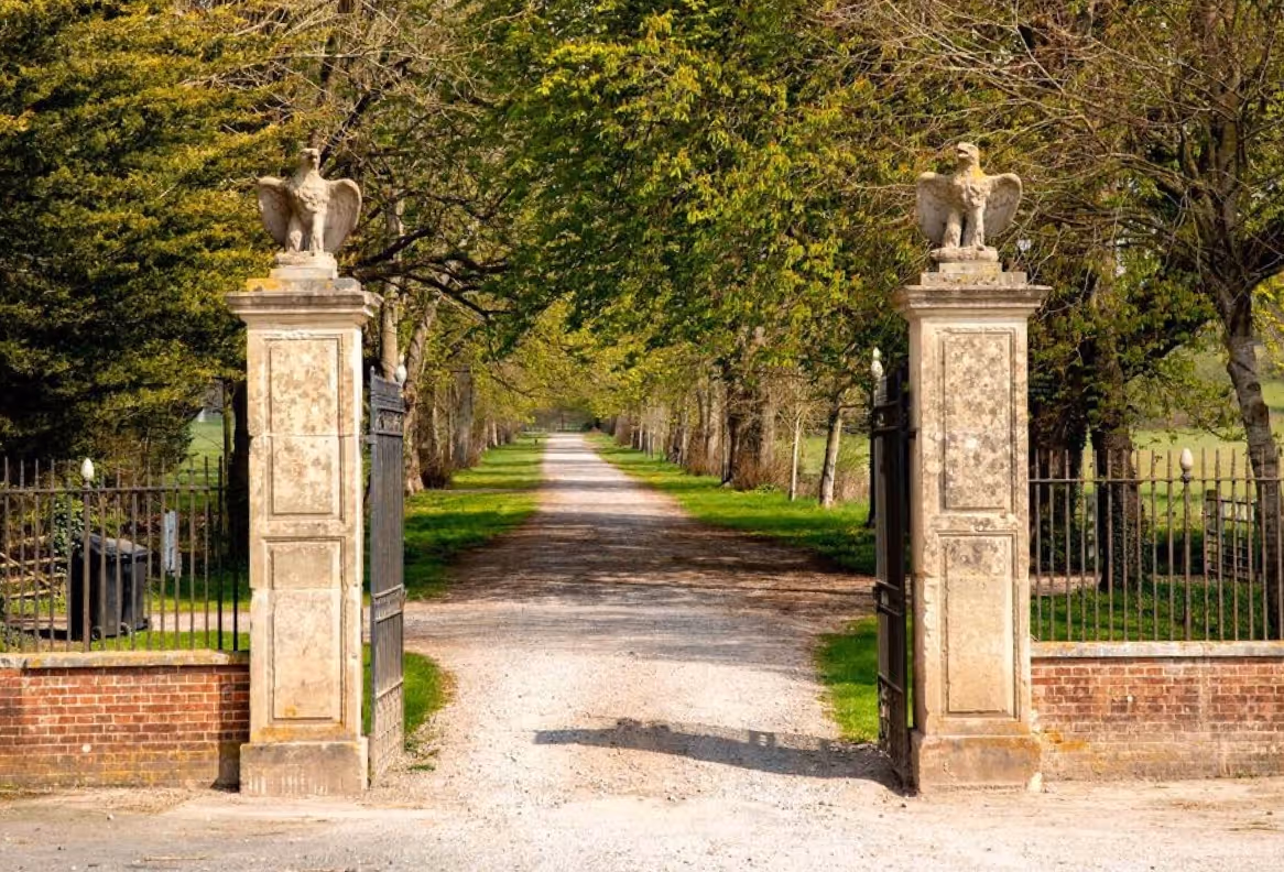 Open wrought iron gates between two stone pillars topped with eagle statues, leading to a tree-lined gravel driveway.