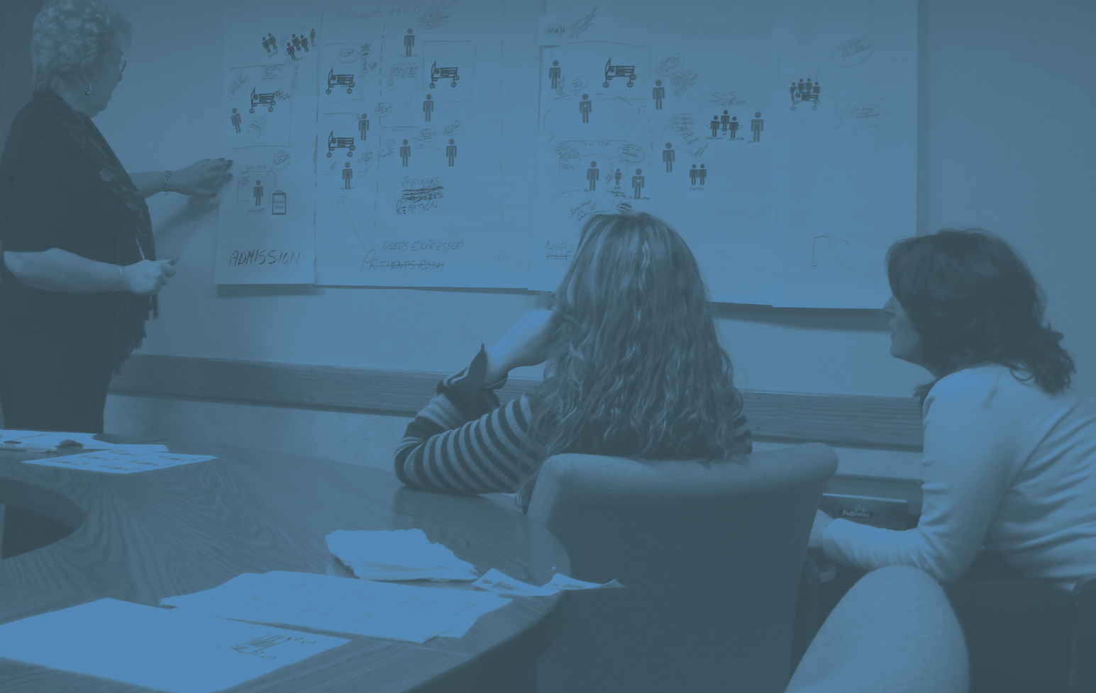 A woman standing and pointing at diagrams on a wall while two seated women attentively watch during a meeting.
