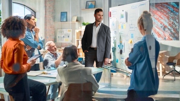 A diverse group of five professionals engaged in a meeting with one man standing and explaining ideas on a whiteboard in a modern office.