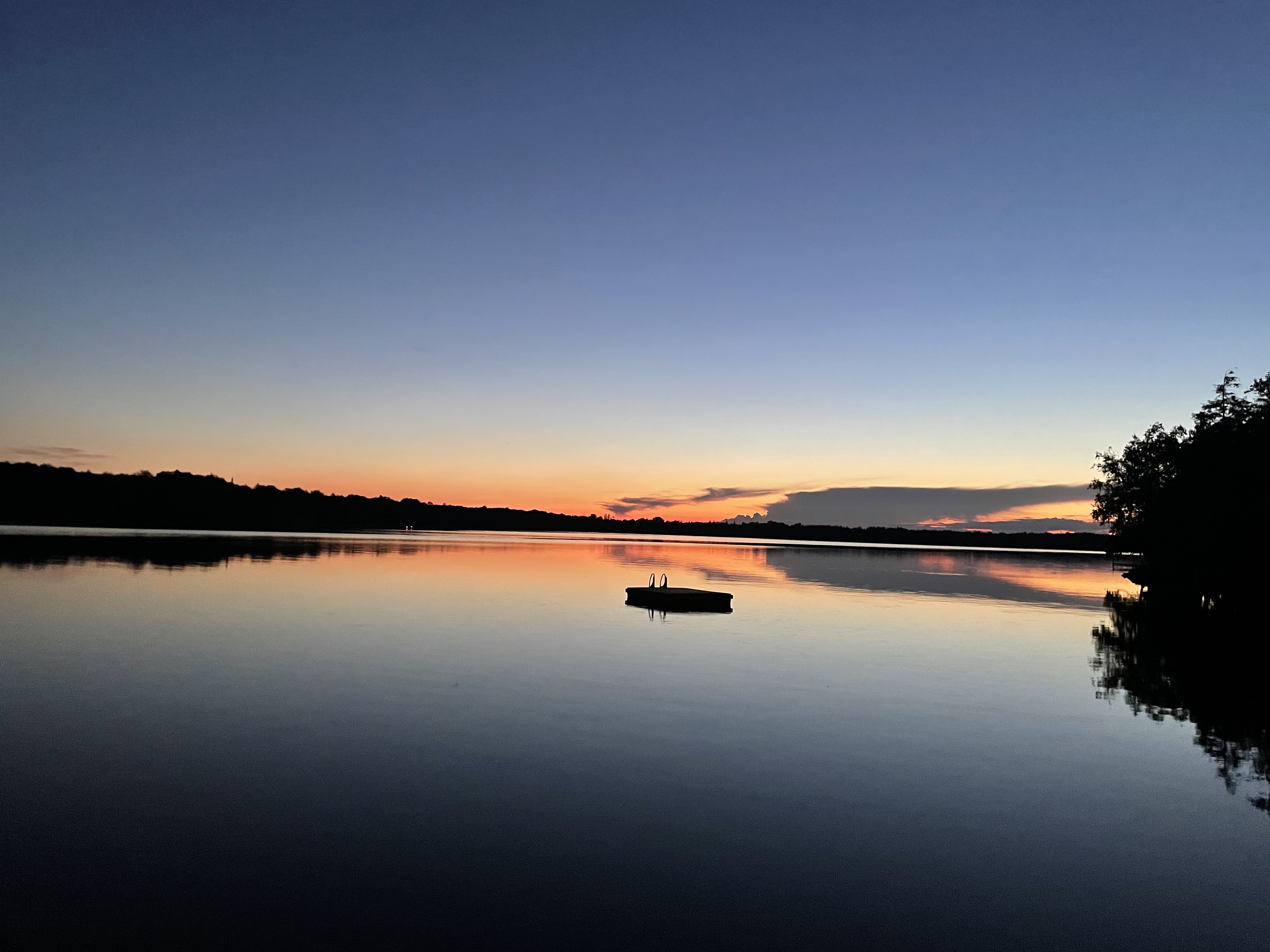 Calm lake at sunset with a small floating dock and silhouette of trees on the horizon.
