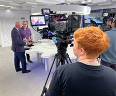 Cameraperson filming two men having a discussion in a modern TV studio with technical equipment and screens in the background.