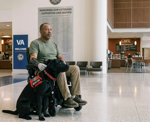 A man in a wheelchair with a prosthetic arm sits indoors accompanied by a black service dog wearing a red vest.