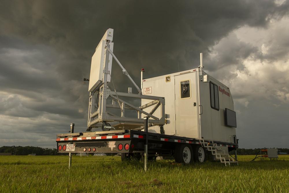 Mobile radar station trailer on grass field under dark, stormy sky with heavy clouds.