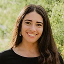 Young woman with long brown hair smiling outdoors with green foliage background