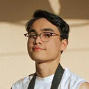Portrait of a young man wearing glasses and a white shirt with a black strap over his shoulder, smiling slightly against a neutral background.