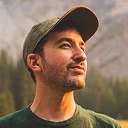 Profile of a bearded man wearing a camouflage cap looking upwards against a blurred outdoor background.
