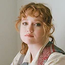 Portrait of a woman with curly light brown hair wearing a patterned jacket and white shirt against a neutral background.