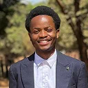 Smiling man wearing a dark blazer and white shirt outdoors with trees in the background.