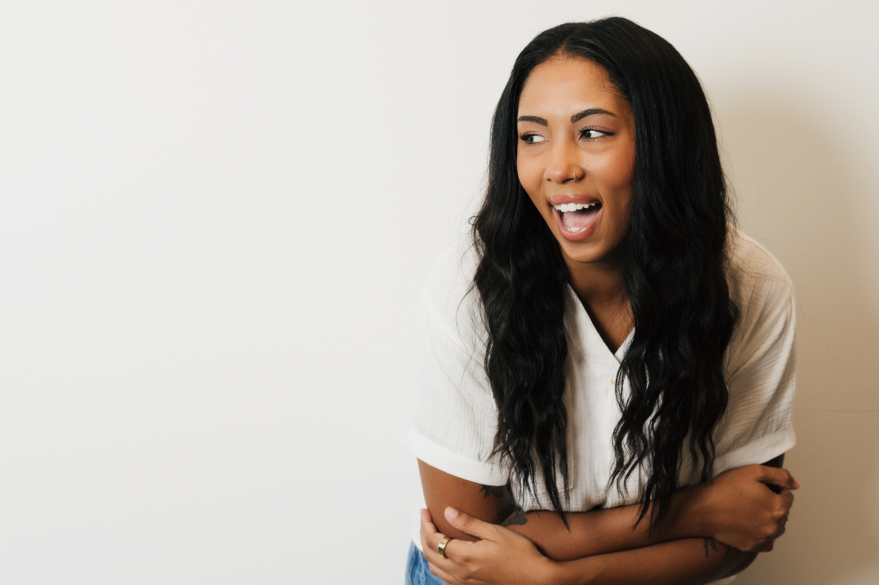Smiling woman with long black hair wearing a white shirt, looking to the side with arms crossed.