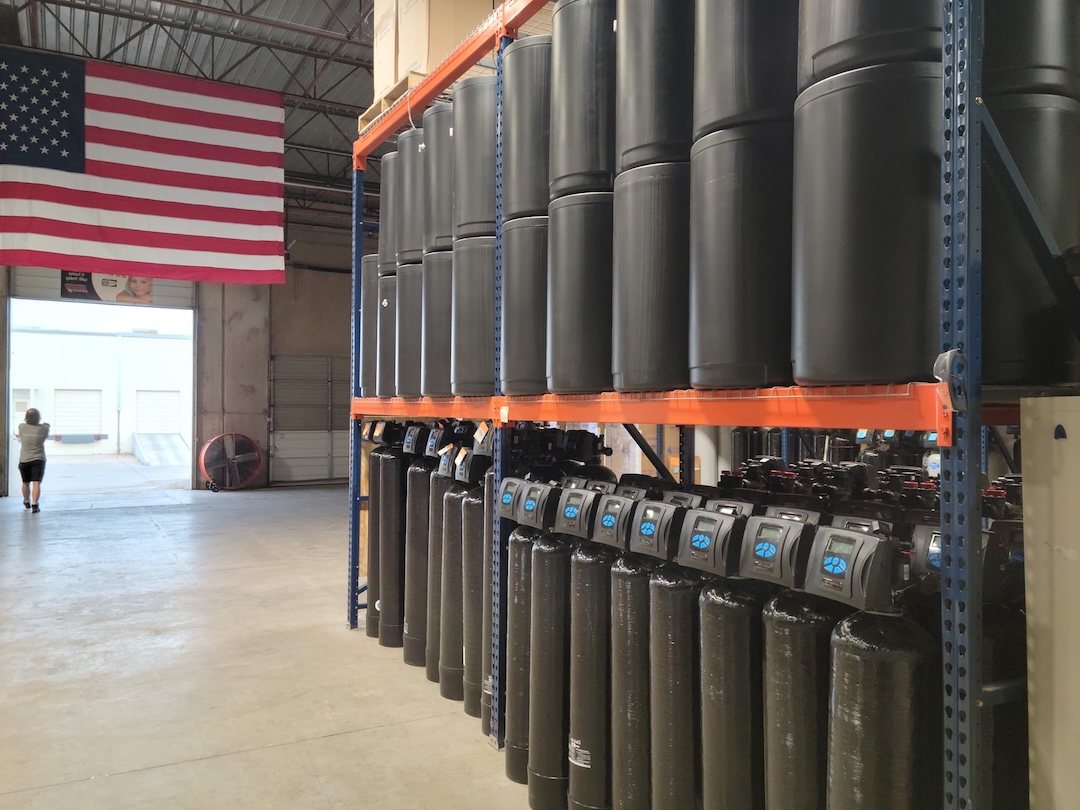 Warehouse interior with tall shelves holding large black cylindrical water filtration tanks and an American flag hanging on the wall.