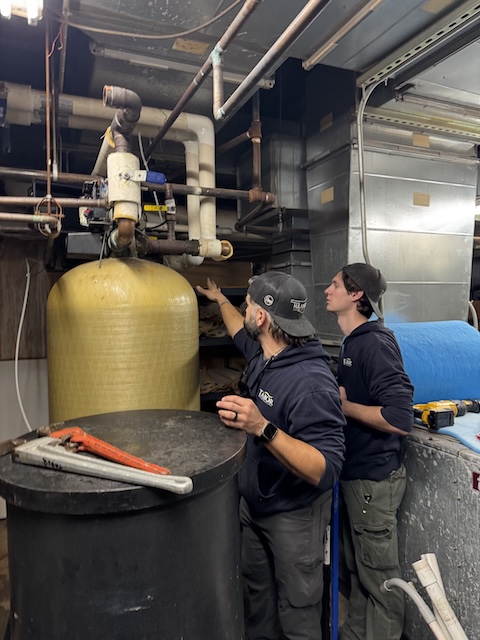 Two workers fitting pipes and equipment in a mechanical room with water filtration tanks.