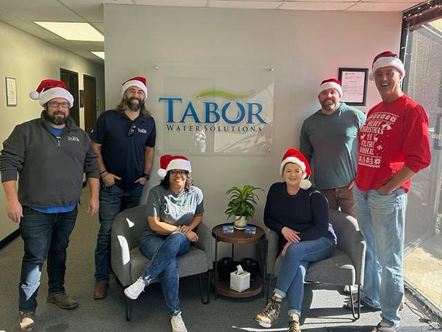 Six people wearing Santa hats posing inside an office with Tabor Water Solutions sign on the wall.