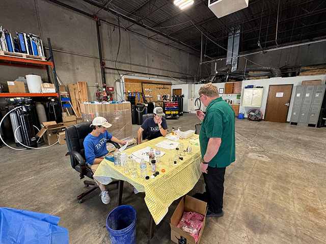 Three men in a large warehouse lab setting examine samples on a table covered with a yellow checkered cloth.