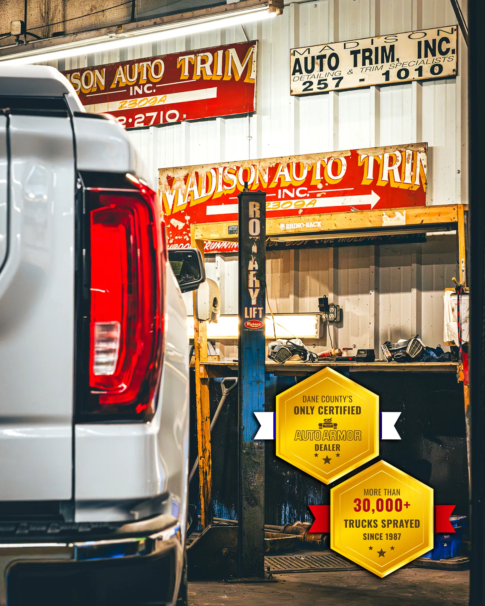 Photo of a white pickup truck stationed on a rustproofing hoist right after a finished professional spray-in bedliner installation. In the background are decade old automotive signs from previous years that read "Madison Auto Trim Since 1987"