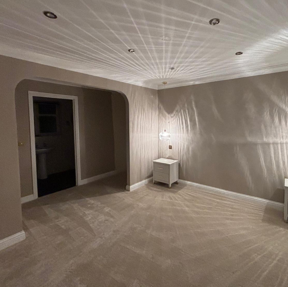 Empty beige carpeted room with a white nightstand and a hanging light casting patterned shadows on walls and ceiling.