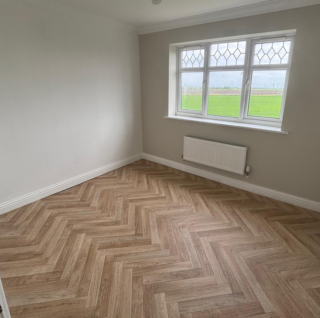Empty room with beige walls, herringbone-patterned wooden floor, large window with diamond glass panes, and white radiator.