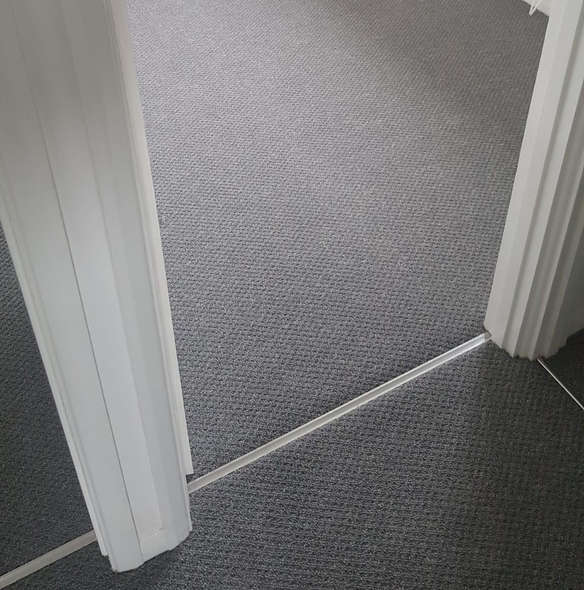 Two adjoining rooms with grey textured carpet flooring separated by a white door frame and a silver metal strip.
