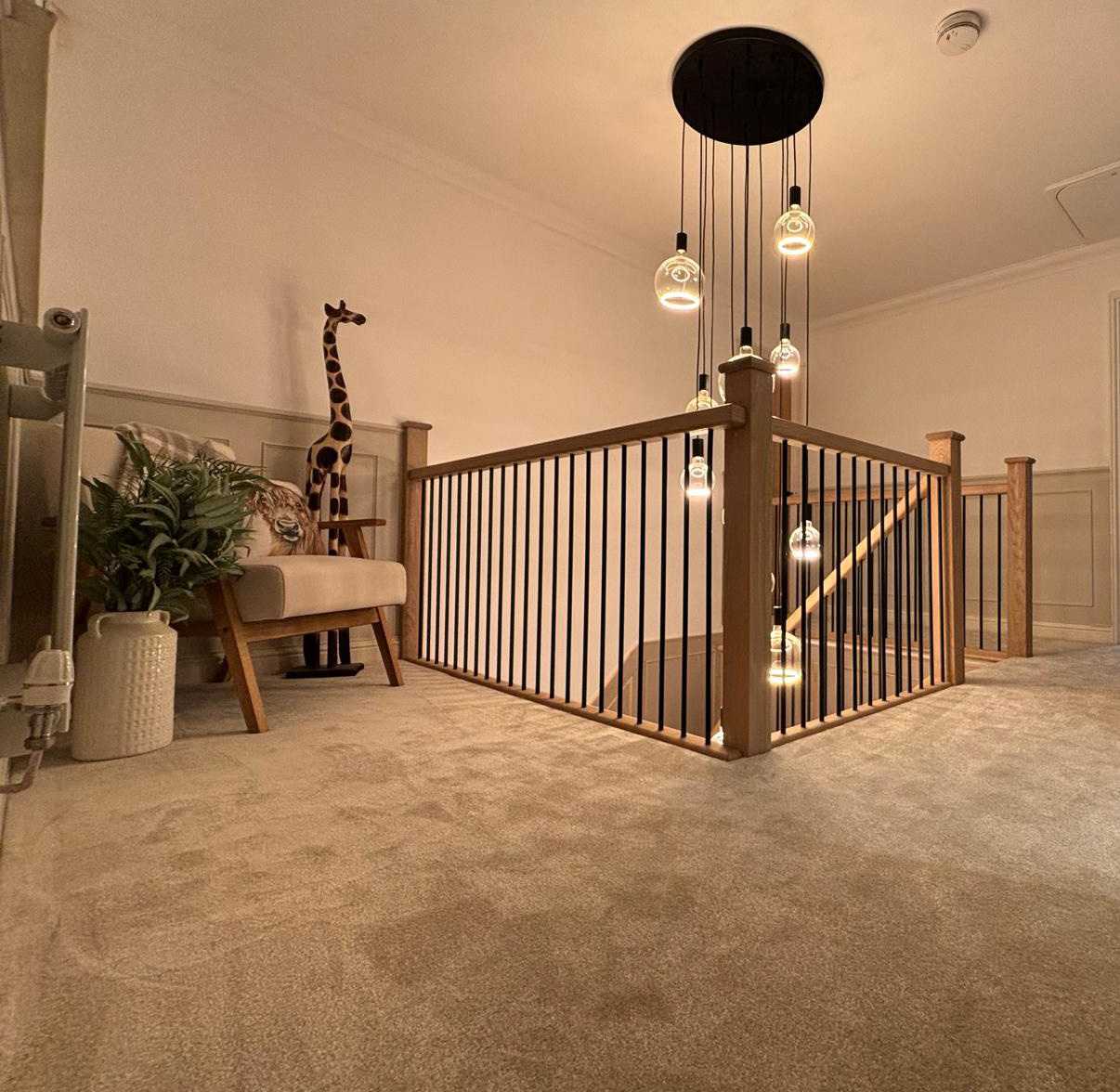 Carpeted upstairs hallway with wooden railing, modern hanging pendant lights, a beige armchair with a tiger cushion, a tall giraffe sculpture, and a plant in a white vase.