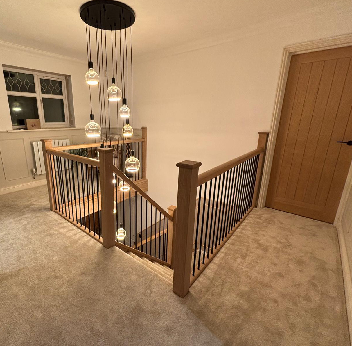 Carpeted upstairs hallway with wooden and black metal railing around staircase and modern hanging pendant lights.