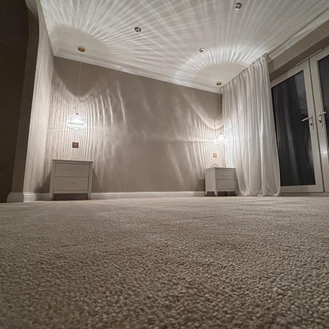 Room with beige carpeted floor, two white nightstands each with a hanging light above, and white curtains by glass doors.