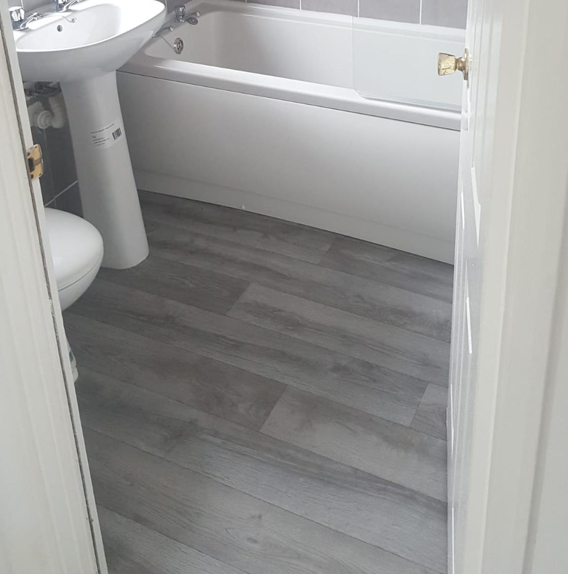Bathroom interior with grey wood-look vinyl flooring, white sink, toilet, and bathtub.