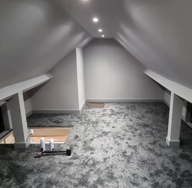 Empty attic room with sloped ceiling, gray walls, recessed ceiling lights, and newly installed gray carpet with some tools on the floor.