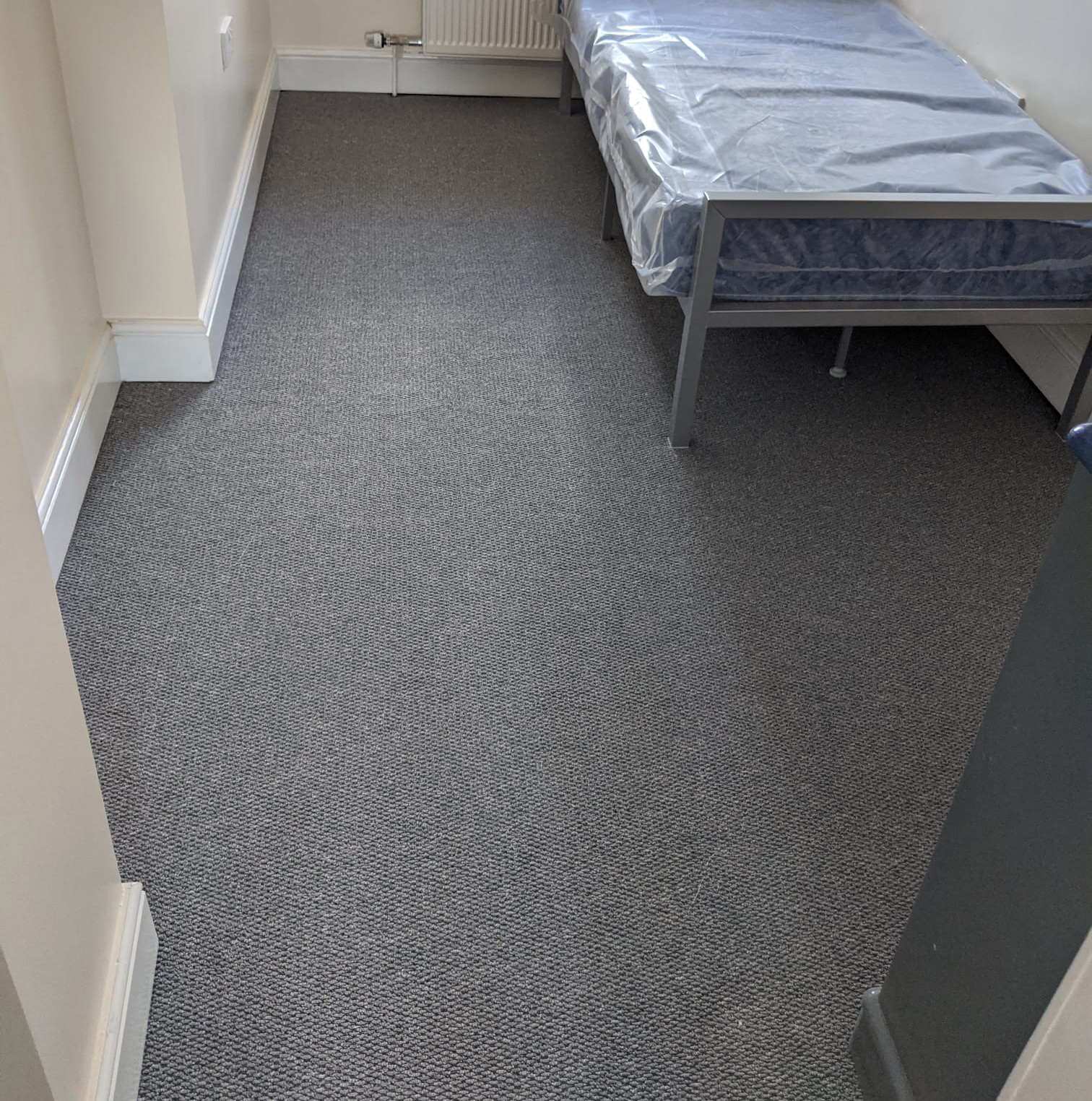 Small bedroom with textured gray carpet flooring and a metal-framed bed covered in plastic.