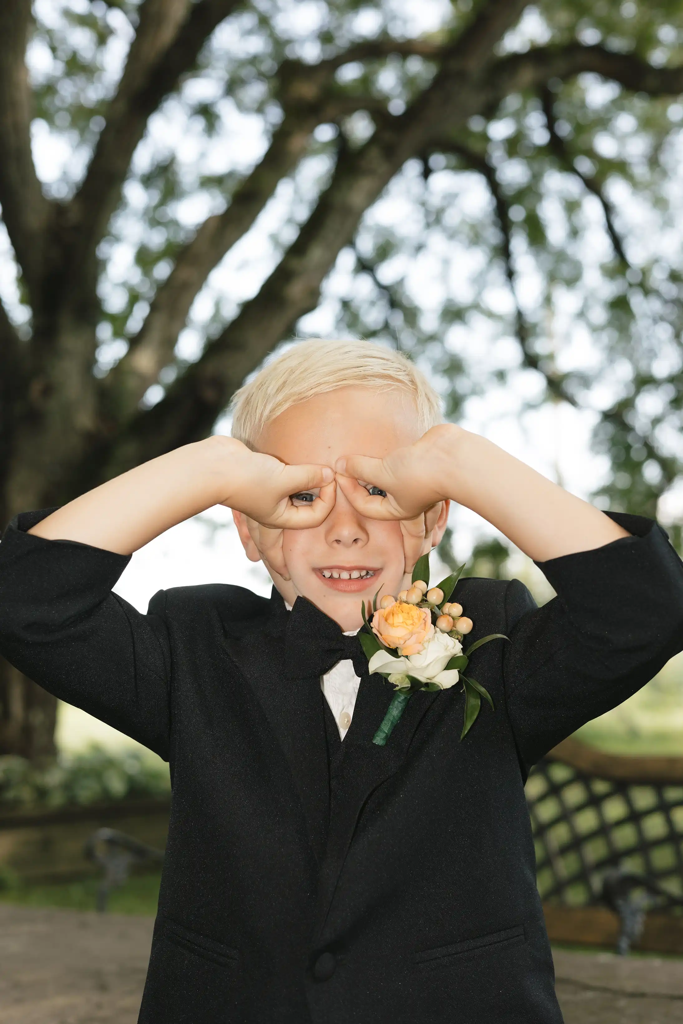 Young boy in a black suit and bow tie with a boutonniere playfully making binoculars with his hands over his eyes.