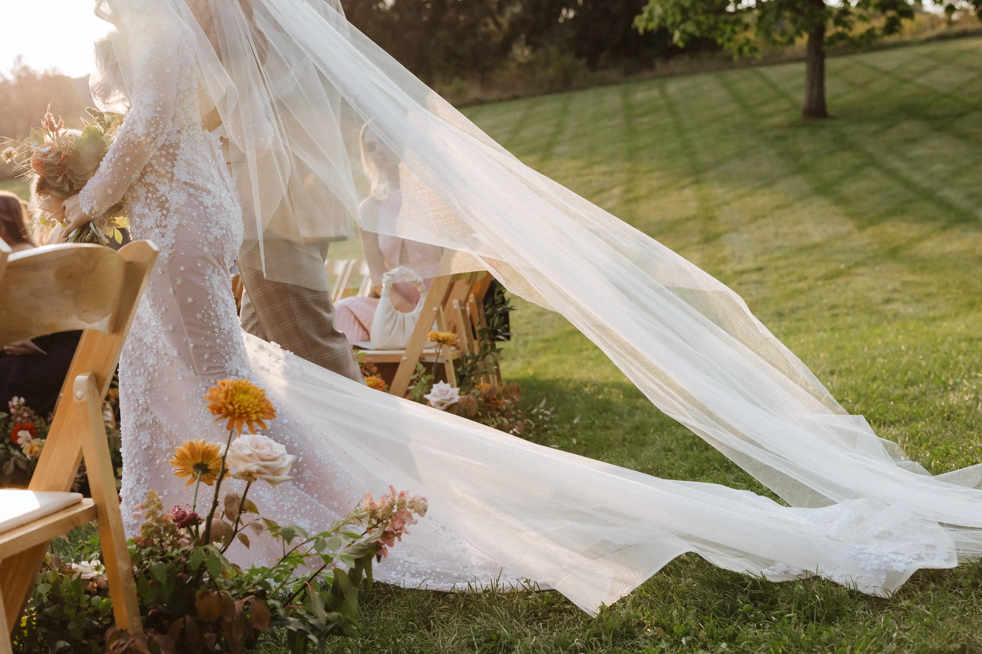 Bride in a white lace wedding dress with a long flowing veil walking outdoors past wooden chairs and flowers on green grass.