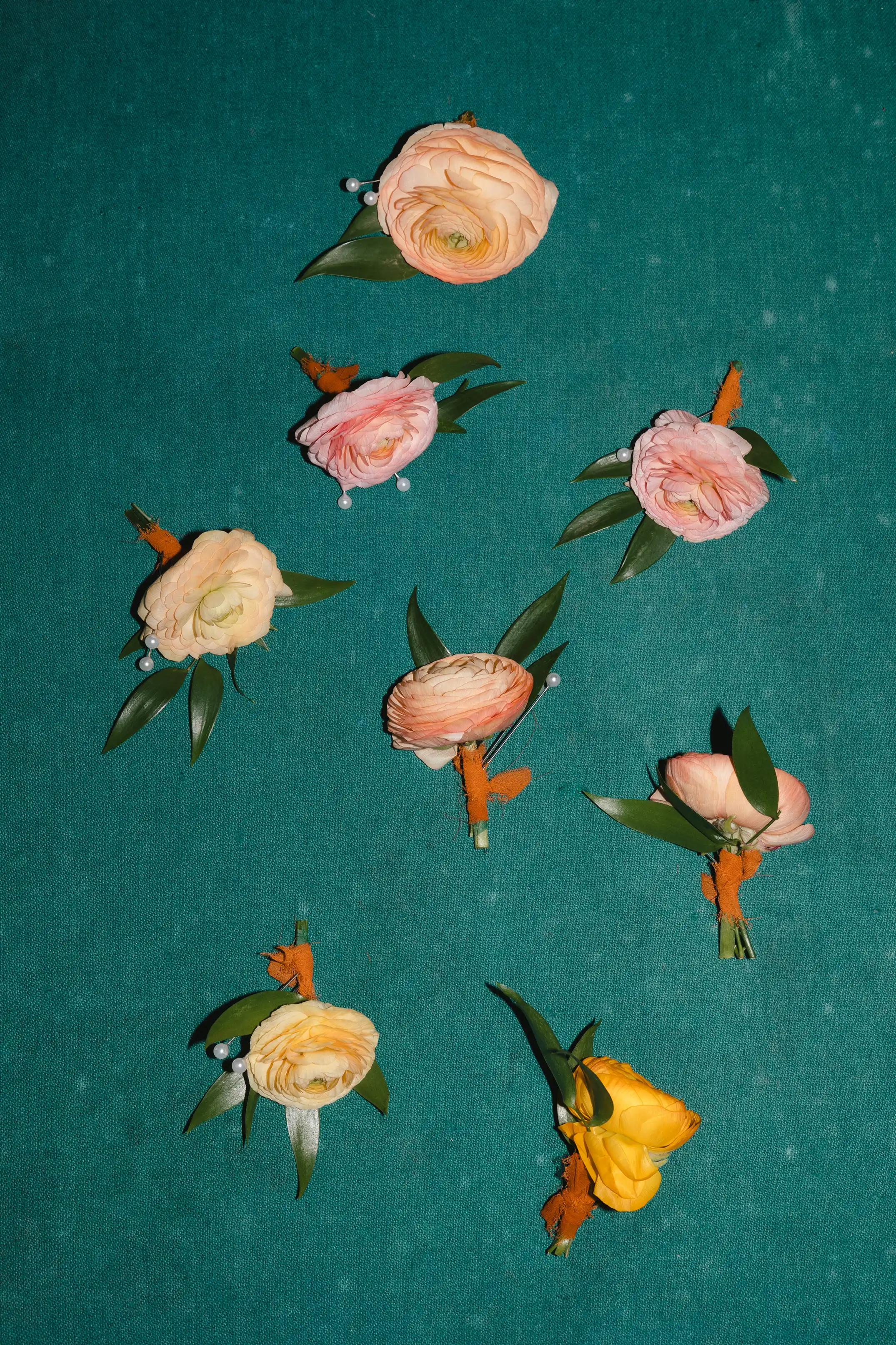 Seven boutonnières made of peach and yellow ranunculus flowers with green leaves and orange floral tape on a teal fabric background.