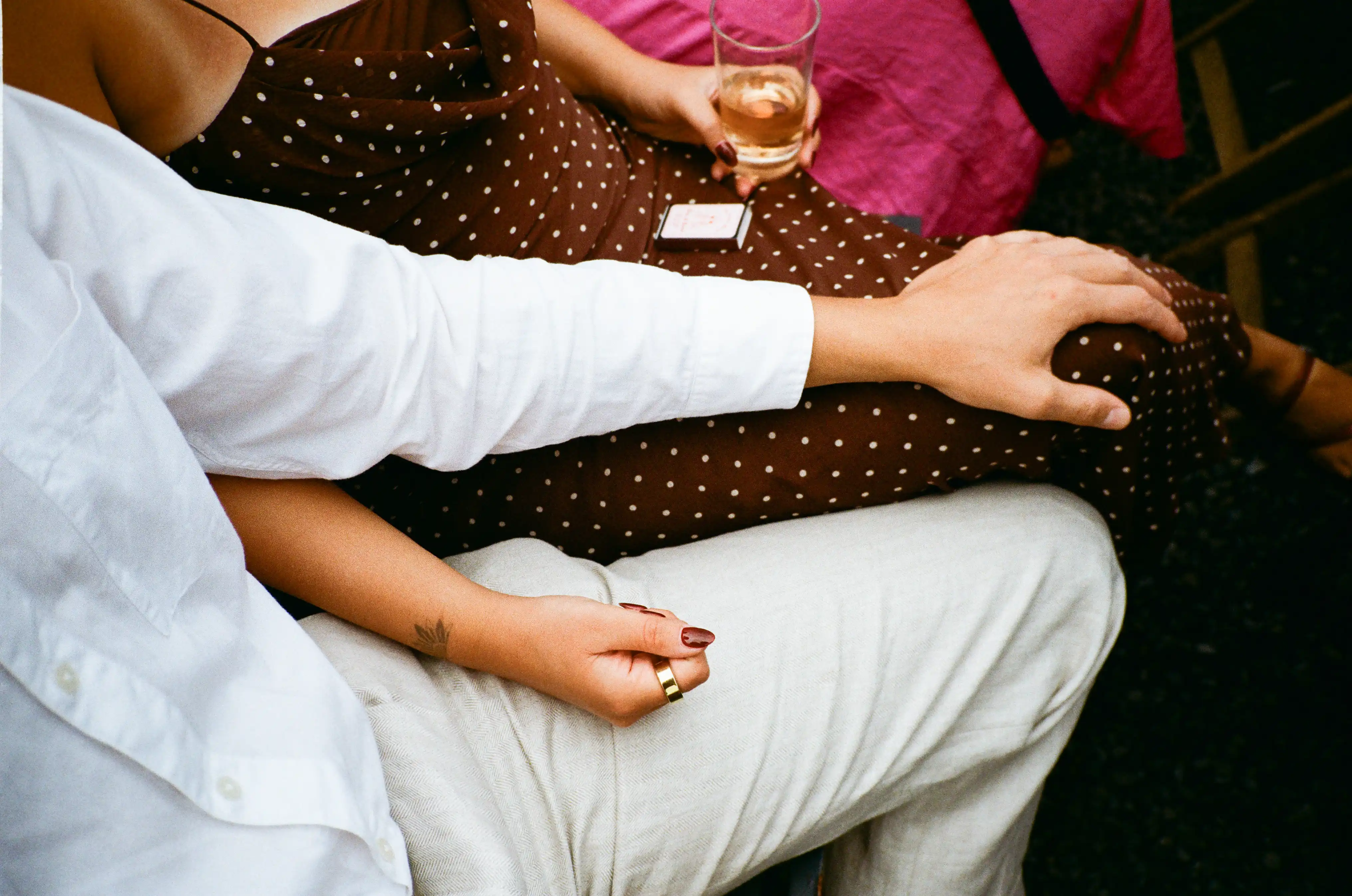Close-up of a couple sitting closely with one person wearing a white shirt and light pants, the other in a brown polka dot dress holding a glass.