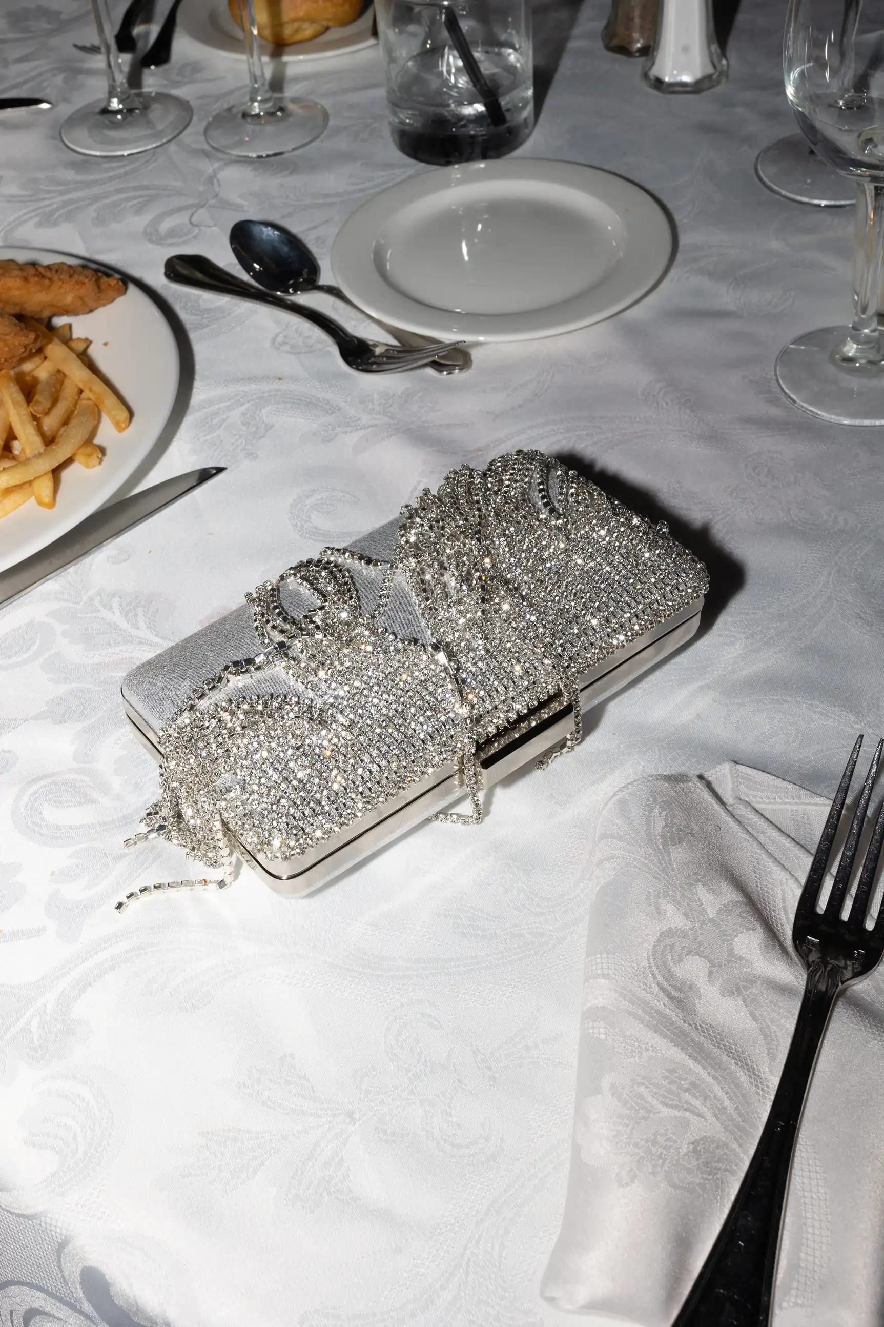 Silver rectangular clutch purse adorned with sparkling rhinestones placed on a white tablecloth near dinnerware and a plate of fries and chicken tenders.