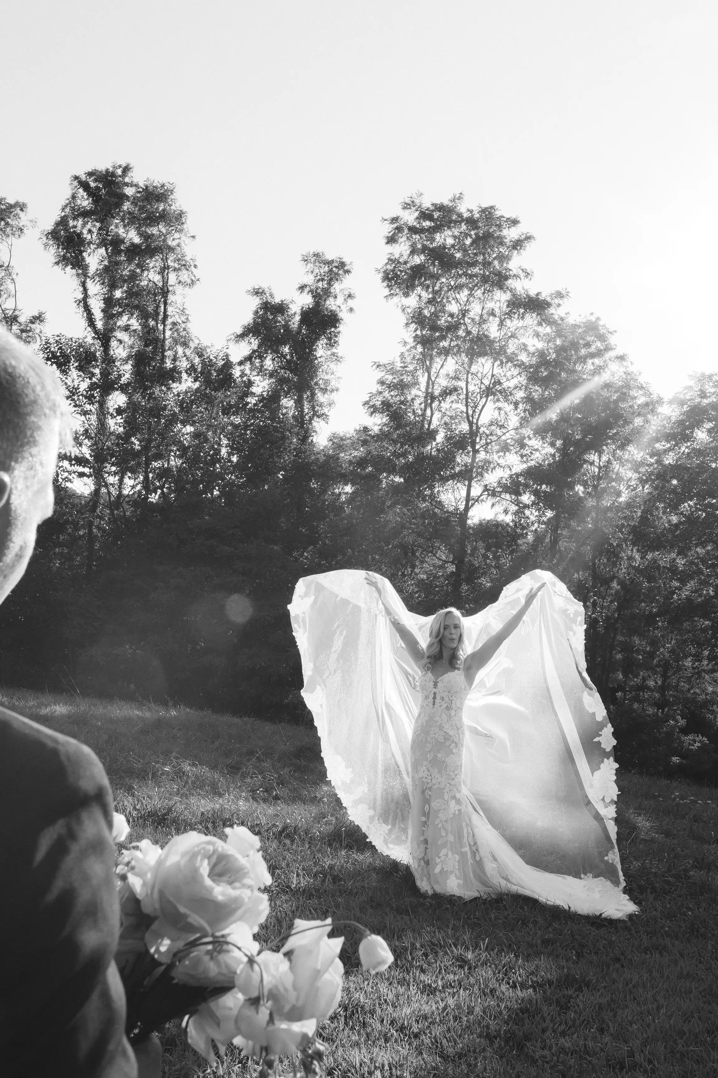 Bride in a lace wedding dress holding out her veil like wings outdoors with a groom holding a bouquet in foreground, black and white photo.