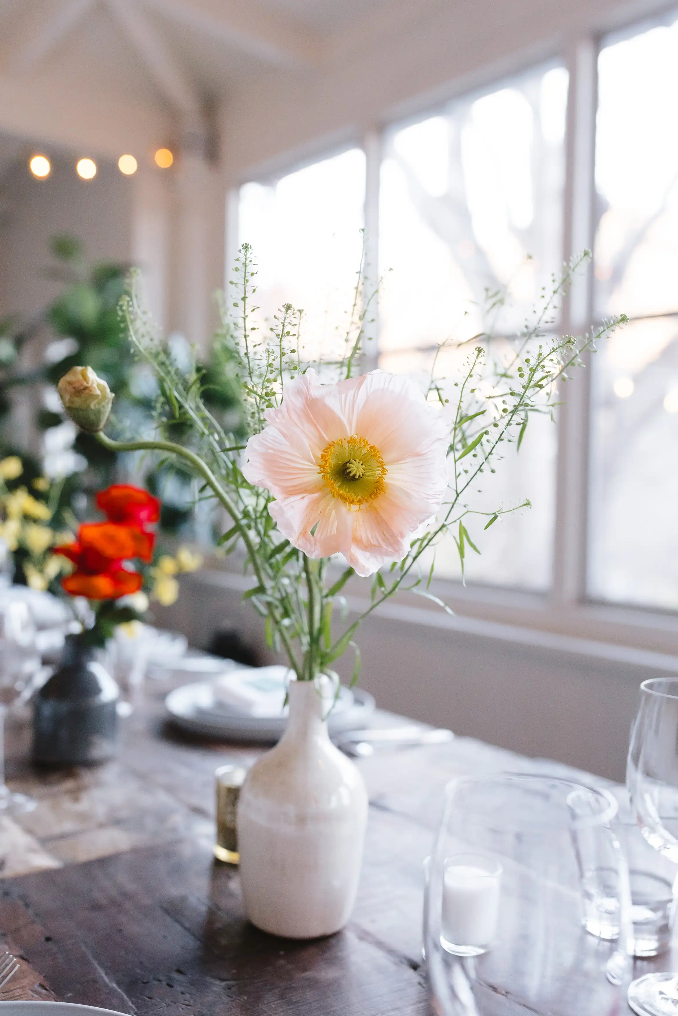 White ceramic vase with a pale pink flower and greenery on a wooden dining table with blurred red flowers and place settings in the background.