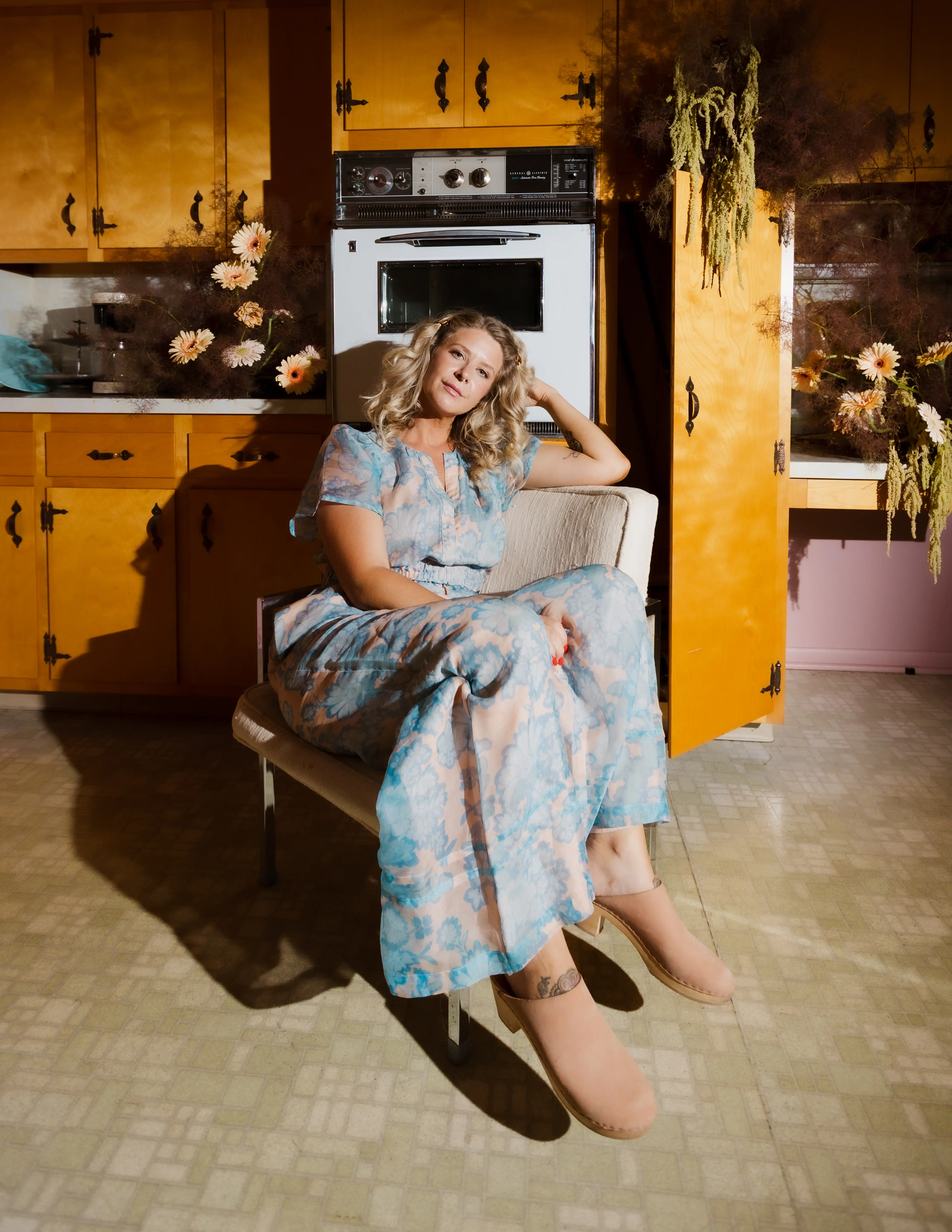 Kathleen Whittemore sits relaxed on a chair in a kitchen with yellow cabinets and dried flowers.